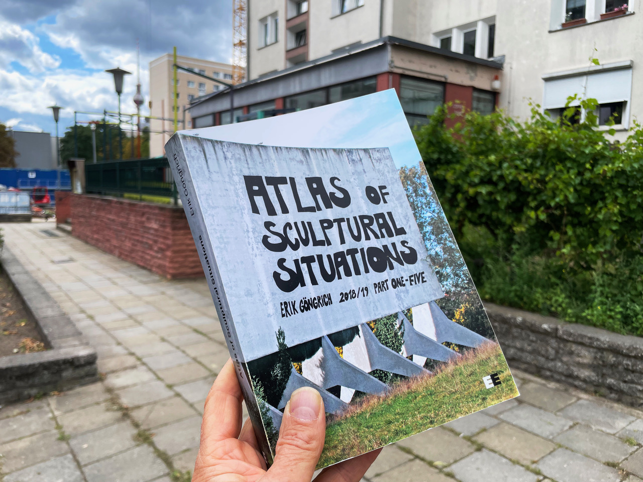 Eine Hand hält das Buch „Atlas of Sculptural Situations“ vor den einem Haus und einem Garten mit Weinblättern