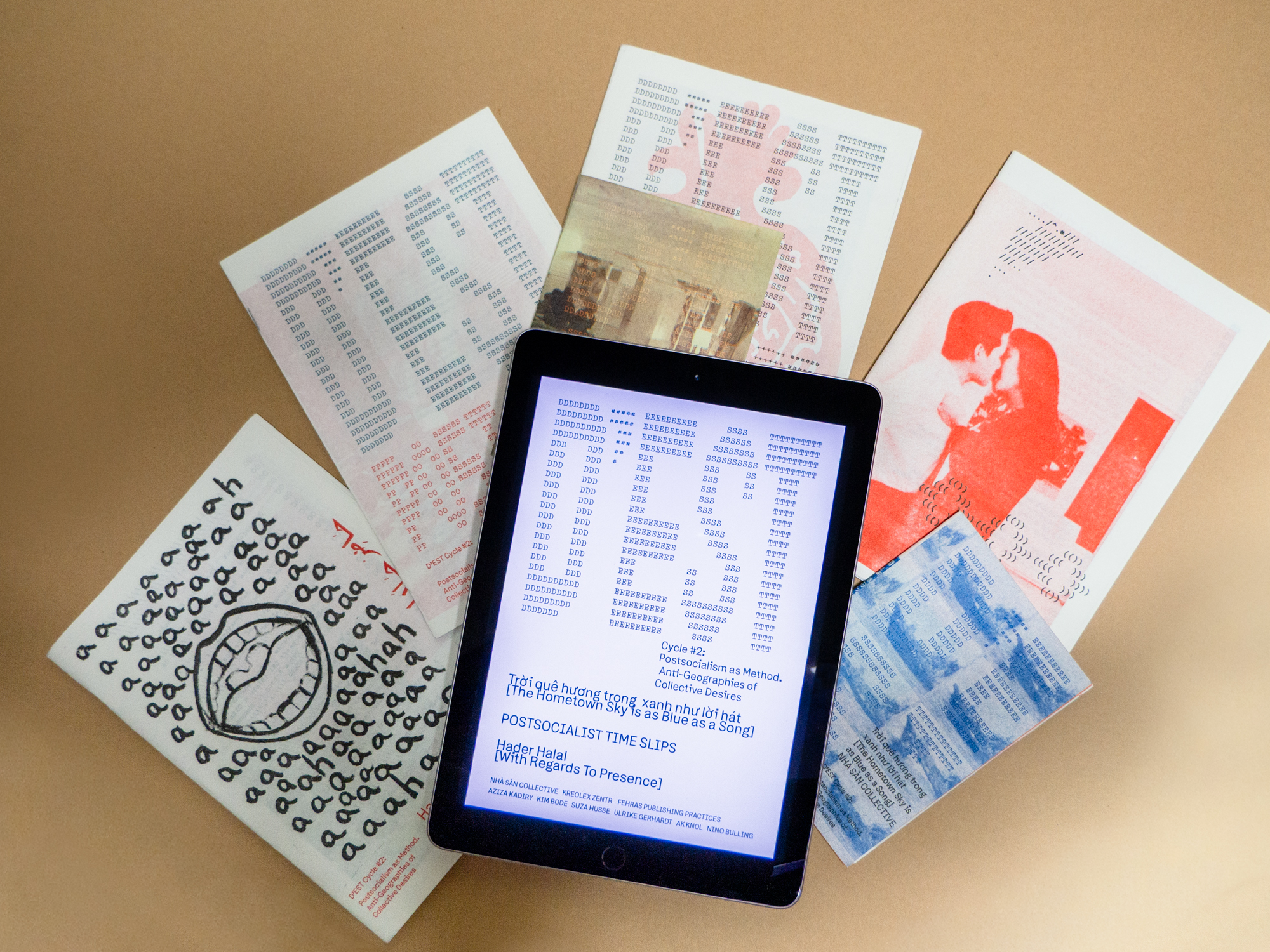 Fünf verschiedene Zines mit unterschiedlichen abstrakten Covern auf beigem Hintergrund; in der Mitte ein iPad, auf dem das Cover der E-Book-Reihe „POSTOST“ zu sehen ist.